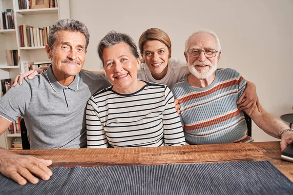 Caregiver looking at camera and smiling while embracing with group of old people
