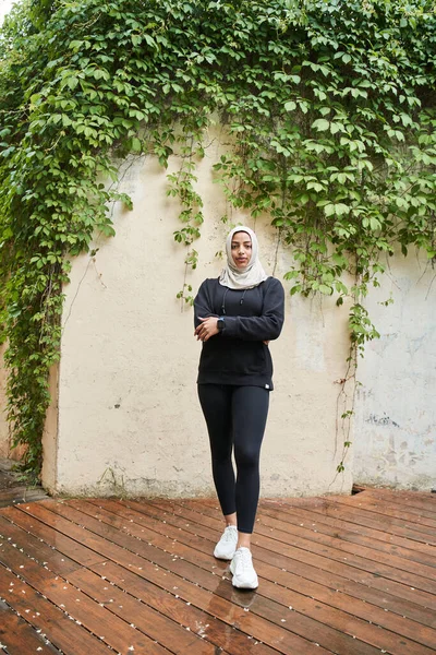 Mujer en hijab sonriendo mientras se prepara para correr en la calle durante el día lluvioso —  Fotos de Stock