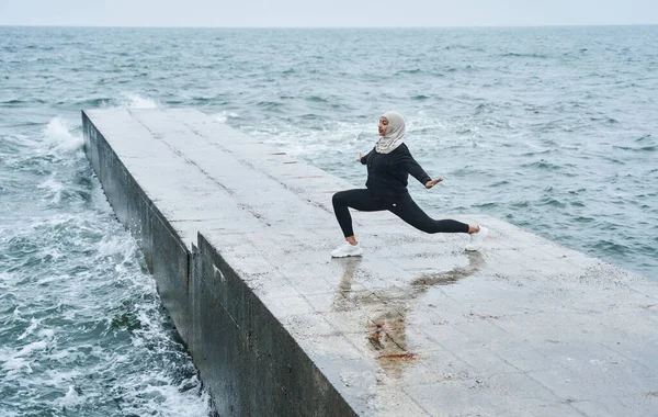 Woman dressed in hijab and dark clothes posing while making sport stretching exercises