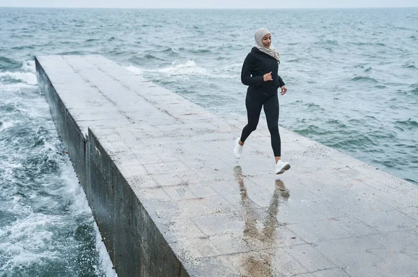 Woman dressed in hijab and dark clothes running outdoors through the pier