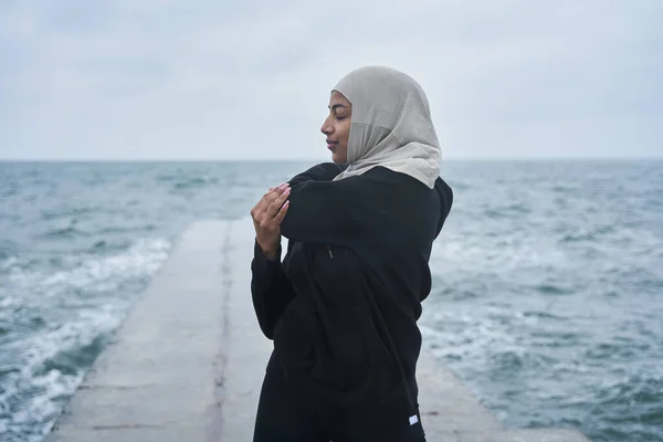 Mujer vestida con hijab y ropa oscura posando mientras hace ejercicios de estiramiento deportivo —  Fotos de Stock