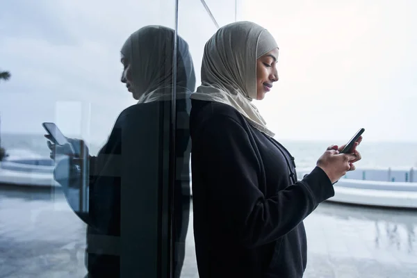 Pani w hidżabie trzyma telefon komórkowy i patrzy na ekran z uśmiechem — Zdjęcie stockowe