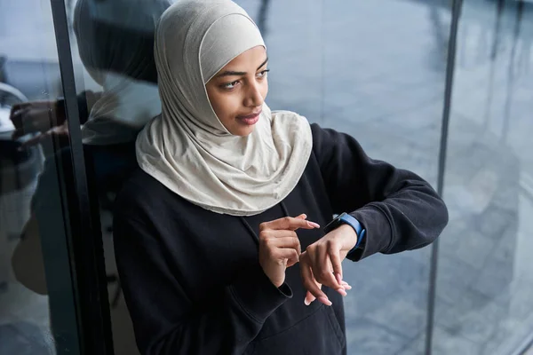 Mujer en el tiempo de seguimiento de hijab en smartwatch mientras está de pie en la calle cerca de las nubes —  Fotos de Stock