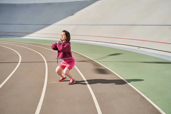 Midget woman wearing sports wear exercising in outdoor arena while doing squats