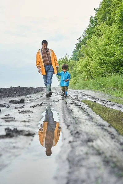 Мальчик идет рука об руку со своим многонациональным отцом по болотистой дороге — стоковое фото