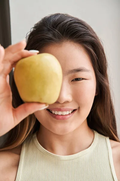 Meisje glimlachend tand en kijkend naar de camera terwijl ze haar oog achter de appel verbergt — Stockfoto