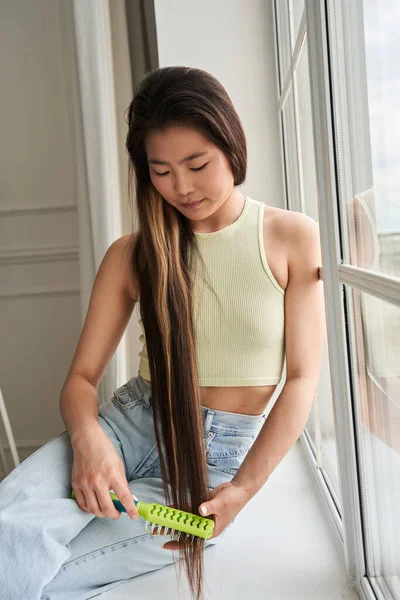 Γυναίκα κοιτάζει τα μακριά μαλλιά της και το βουρτσίζει ενώ κάθεται στο περβάζι του παραθύρου — Φωτογραφία Αρχείου