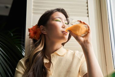 Saçında çiçekle meyve suyu içip pencereye bakan bir kadın.