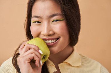 Asyalı kadın yeşil elmayı tutarken hafif bir gülümsemeyle kameraya bakıyor.