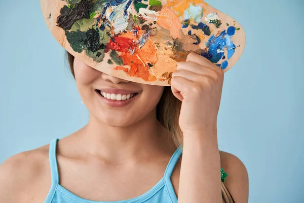 Mujer escondiendo su cara detrás de la paleta con pinturas mientras filma en el estudio —  Fotos de Stock
