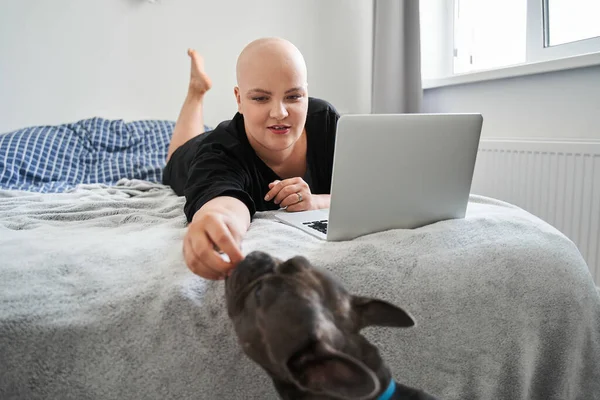 Mujer calva alimentando a su perro mientras está acostada en la cama y trabajando — Foto de Stock