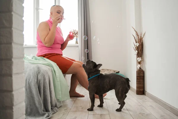 Plešatý žena fouká bubliny s radostí, zatímco tráví čas se svým psem — Stock fotografie
