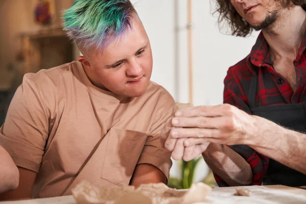 Rzemieślnik w fartuchu uczy młodego chłopca z zespołem Downa robienia talerza z gliny — Zdjęcie stockowe