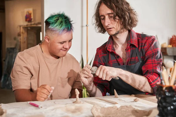 Rzemieślnik pokazując swojemu uczniowi z zespołem Downa, jak rzeźbić z gliny — Zdjęcie stockowe
