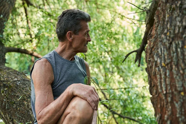 Uomo anziano guardando lontano e godendo della foresta mentre seduto all'albero — Foto Stock