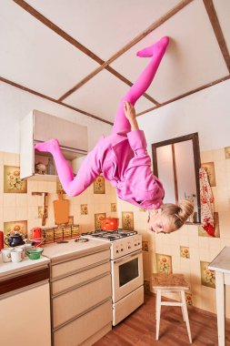 Tavanda tepetaklak duran ve mutfakta vakit geçirirken aşağıya bakan bir kadın.