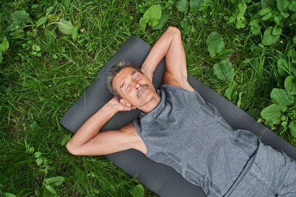 Зрелый человек делает расслабляющую позу йоги, лежа на коврике на природе — стоковое фото