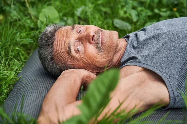 Старший мужчина лежит на траве на коврике для йоги и наслаждается природой — стоковое фото