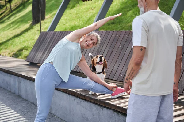 Женщина делает склоны, держа ногу на скамейке рядом с собакой — стоковое фото