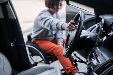 Woman in wheelchair holding at the steering wheel and getting into the car on city street