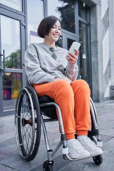 Dziewczyna z upośledzeniem dolnej części ciała siedzi na wózku inwalidzkim i patrzy na smartfona — Zdjęcie stockowe