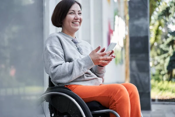 Mujer discapacitada sosteniendo teléfono inteligente y mirando hacia otro lado mientras está sentada en la silla de ruedas — Foto de Stock