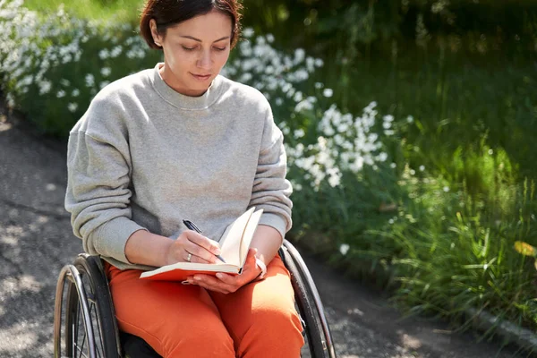 Calm woman sitting at the wheelchair and writing something at her notebook