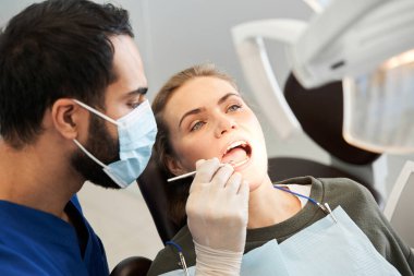 Sarışın bayan müşterisinin yanında duran koruyucu maskeli bir dişçi.