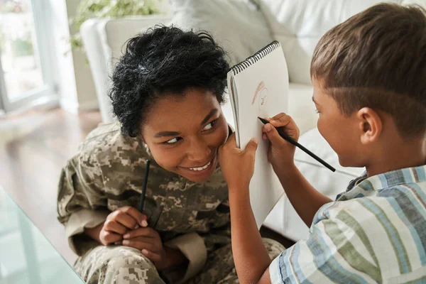 Mujer vistiendo uniforme militar sonriendo y mirando a su hijo mientras él la dibuja — Foto de Stock