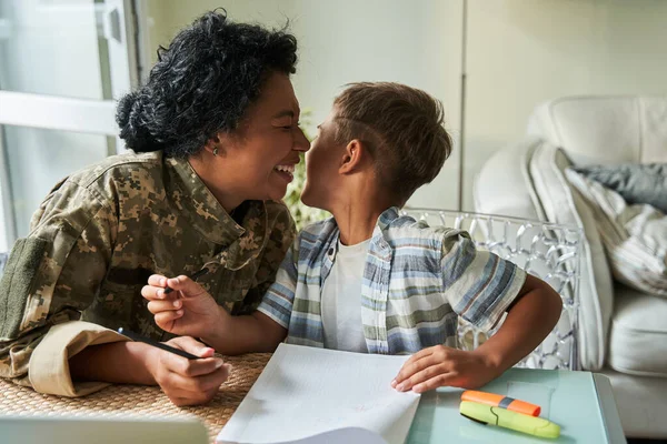 Mujer soldado riendo en voz alta mientras dibuja en la mesa con su pequeño hijo — Foto de Stock