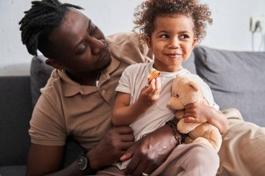 Kanepede otururken küçük kıvırcık kızına bakan bir adam.