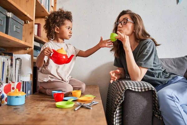 Chica tratando a su madre con el té mientras juega en casa con sus platos de juguete —  Fotos de Stock