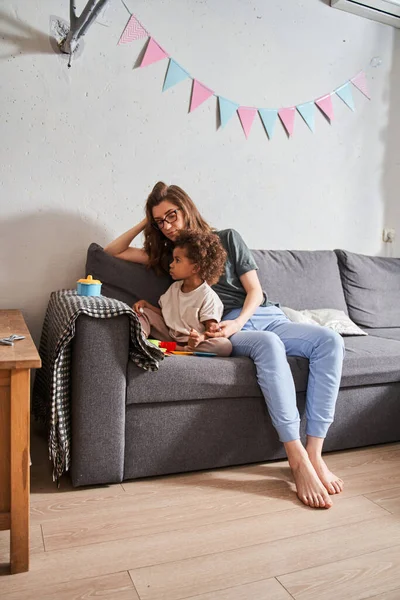 Flicka leker med sina leksaker när hon sitter i soffan nära sin mamma — Stockfoto