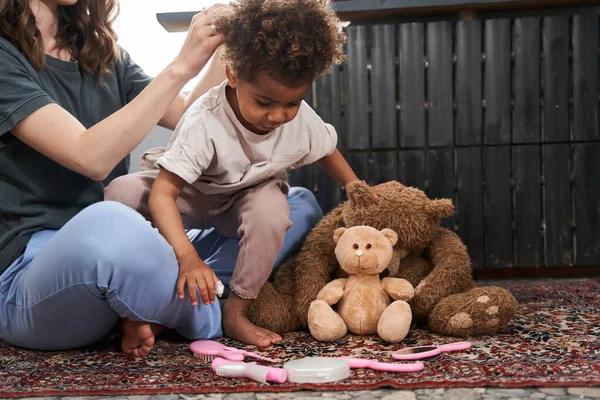 Dívka hraje se svými hračkami, zatímco sedí na podlaze se svou matkou dělá účes — Stock fotografie