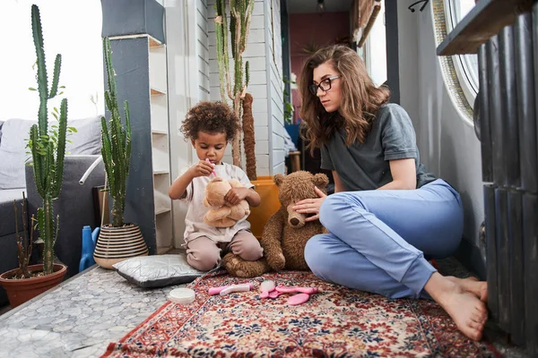 Chica jugando con juguetes de oso de peluche y sonriendo mientras su madre está sentada cerca —  Fotos de Stock