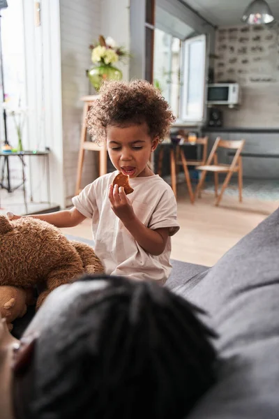 Chica comiendo galleta mientras está sentada en el sofá cerca de su padre y mirándolo con interés — Foto de Stock