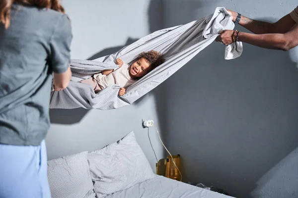 Menina deitado embrulhado no cobertor e rindo em voz alta, enquanto seus pais balançando-a — Fotografia de Stock