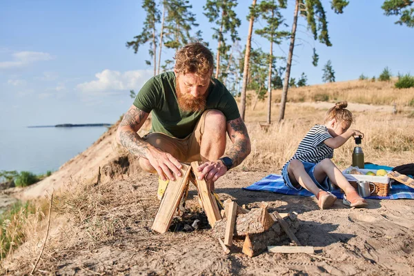 Man making fire from logs on coast near daughter — Stock Photo, Image