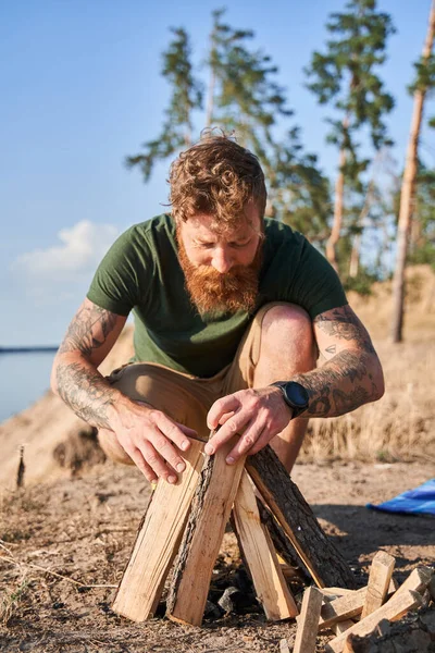 European man making fire from logs on sandy coast — Stock Photo, Image