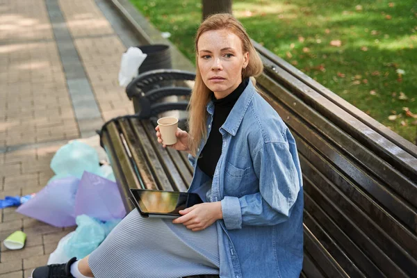 Mujer sentada en el banco alrededor de la basura y usando la tableta mientras bebe café — Foto de Stock