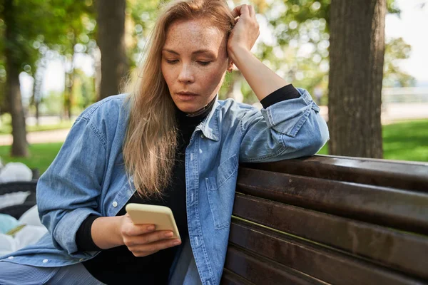 Žena sedící na lavičce na ulici a dívající se na obrazovku chytrého telefonu — Stock fotografie