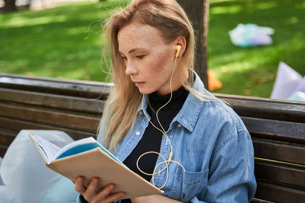 Femme écoutant de la musique aux écouteurs et lisant le livre tout en passant du temps sur le banc — Photo