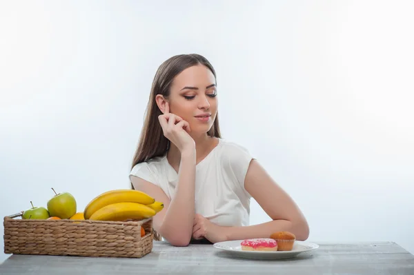 Pretty young woman is choosing between fruits and sweet food — Stock Photo, Image