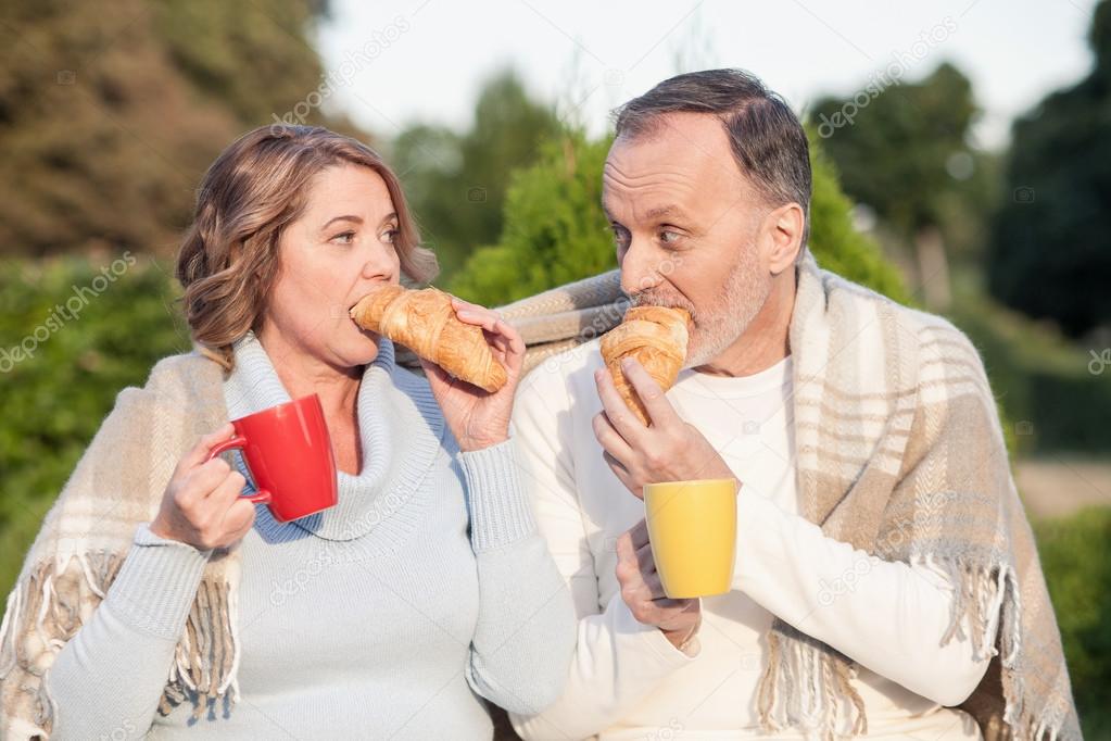 Cheerful senior husband and wife are eating in park