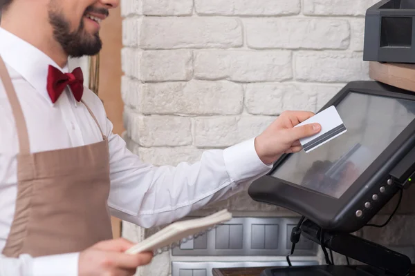 Cheerful young waiter is working with checking machine