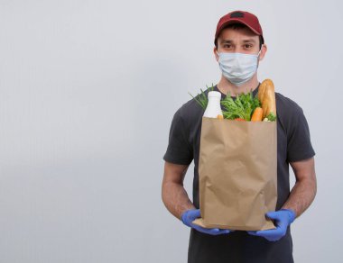 Male courier in a cap, mask and gloves with a paper bag of essentials. Home delivery. Self-isolation delivery concept