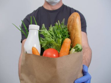 A courier wearing a medical mask and protective gloves delivers online ordering of products. Fresh food vegetables, herbs, milk, loaf in a craft package close-up