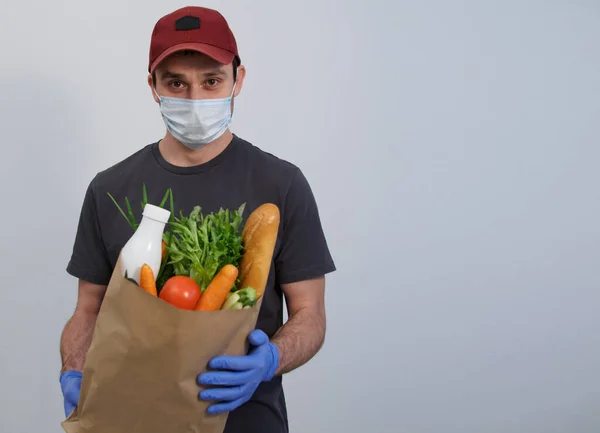 A delivery man in a uniform, mask, gloves and a cap holds a package with groceries on a white background. Mock up copy space