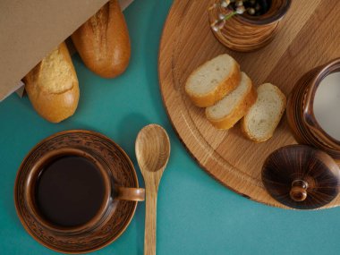 Breakfast in handmade earthenware top view. Cup of tea, milk in a jug, baguette in a paper bag, on a wooden tray pussy willow branches in a vase, wooden spoon on a green background. Flat lay