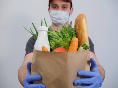 Koruyucu maskeli ve eldivenli bir kurye, arka planda uzanan beyaz bir arka planda içi yiyecek dolu bir kese kağıdı tutuyor. Karantinada eve teslim, salgın hastalık